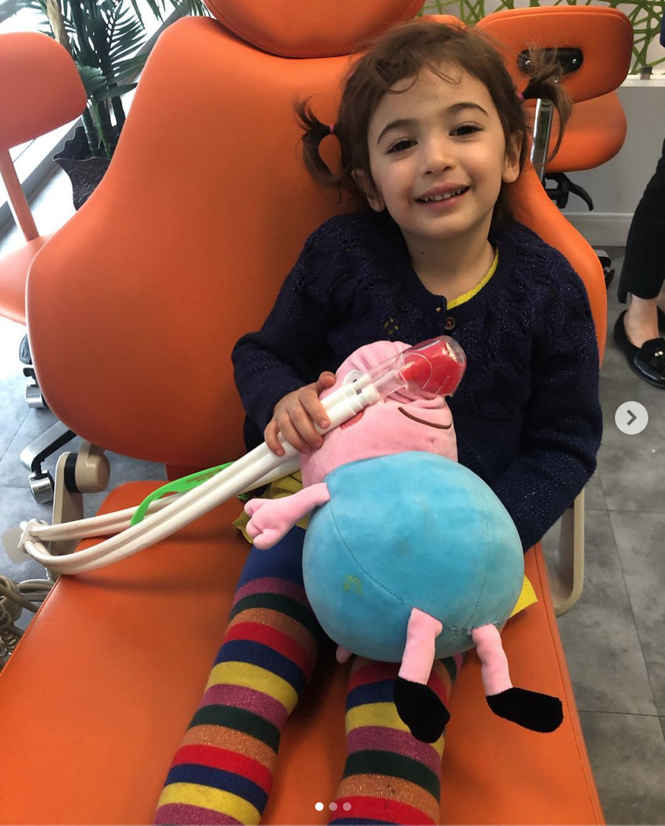Girl with pigtails smiles, holding Peppa Pig toy at dentist office, wearing striped socks and blue sweater.