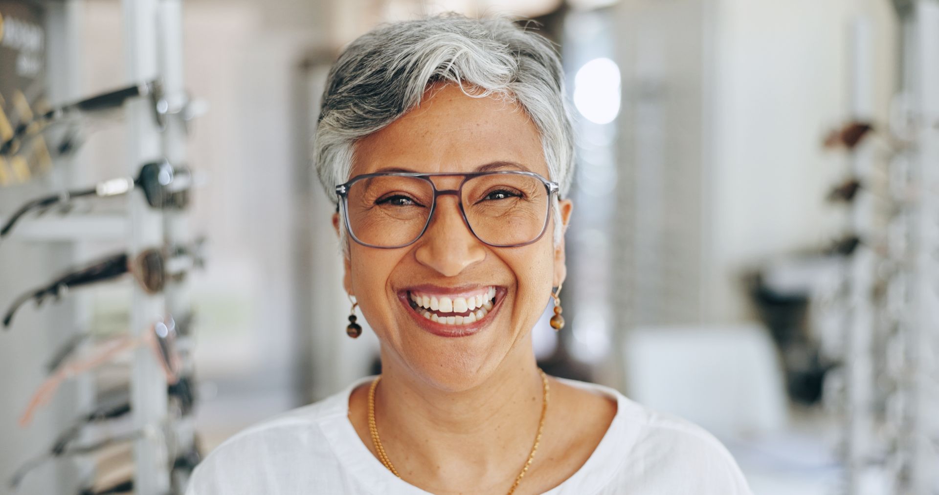 A smiling person with grey hair wears thin-rimmed glasses in an optometry shop with eyewear displays in the background. A smiling person with grey hair wears thin-rimmed glasses in an optometry shop with eyewear displays in the background.