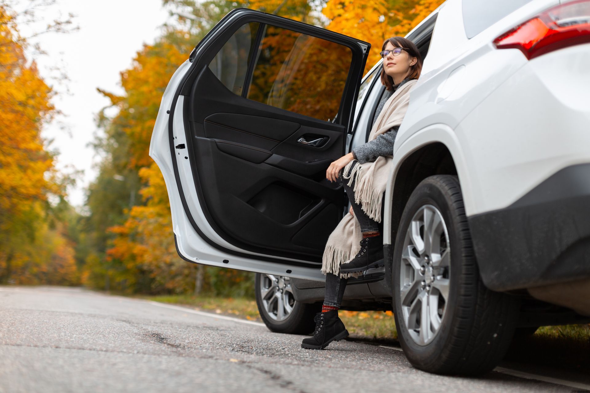 A woman with glasses is sitting in a car doorway with one leg on asphalt on a road in autumn nature. A woman with glasses is sitting in a car doorway with one leg on asphalt on a road in autumn nature.