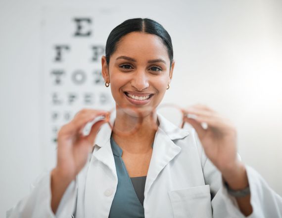 An optometrist in a white coat smiling and holding up a pair of eyeglasses, with an eye chart visible in the background. An optometrist in a white coat smiling and holding up a pair of eyeglasses, with an eye chart visible in the background.