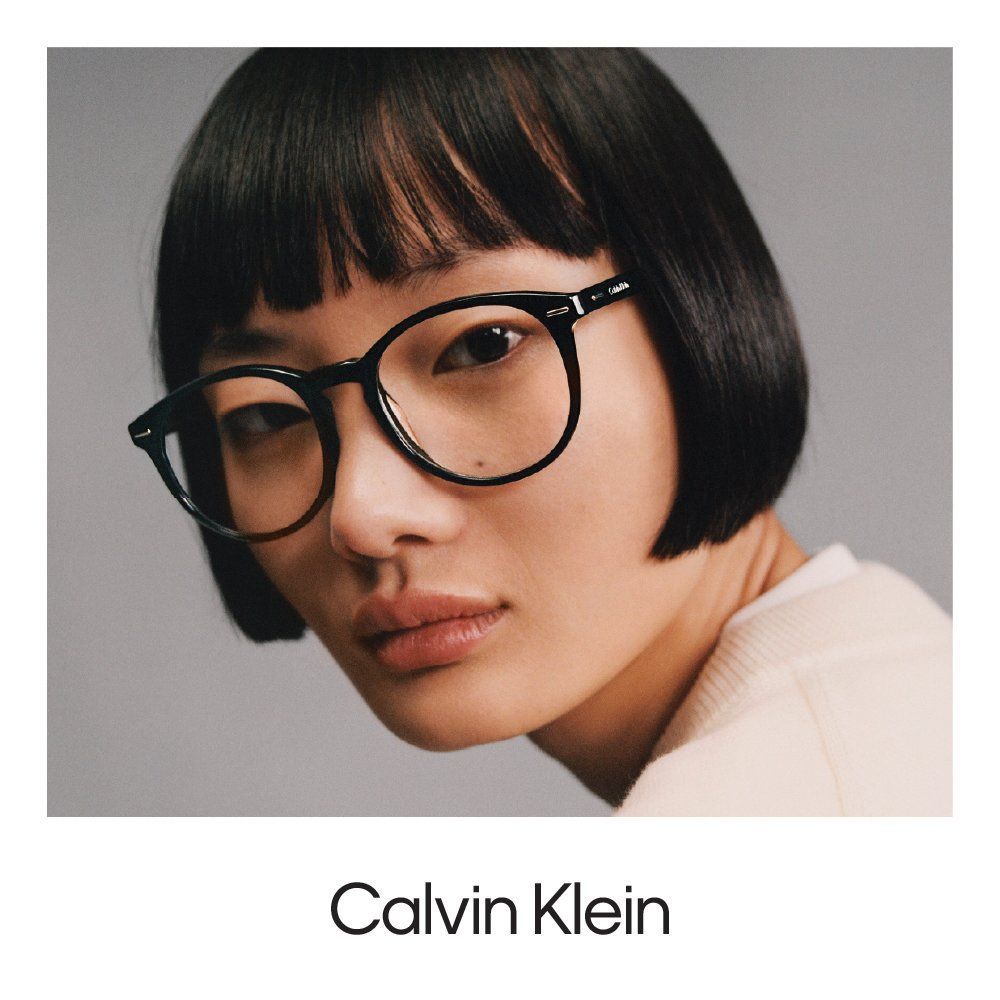 A person with a blunt bob haircut wearing round, black-framed Calvin Klein glasses against a plain gray background.