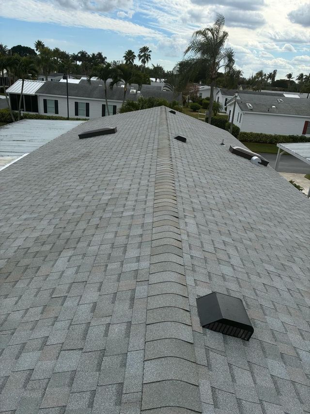 A high-angle view of a gray shingled roof with several black vents and a ridgeline, set in a sunny residential neighborhood.