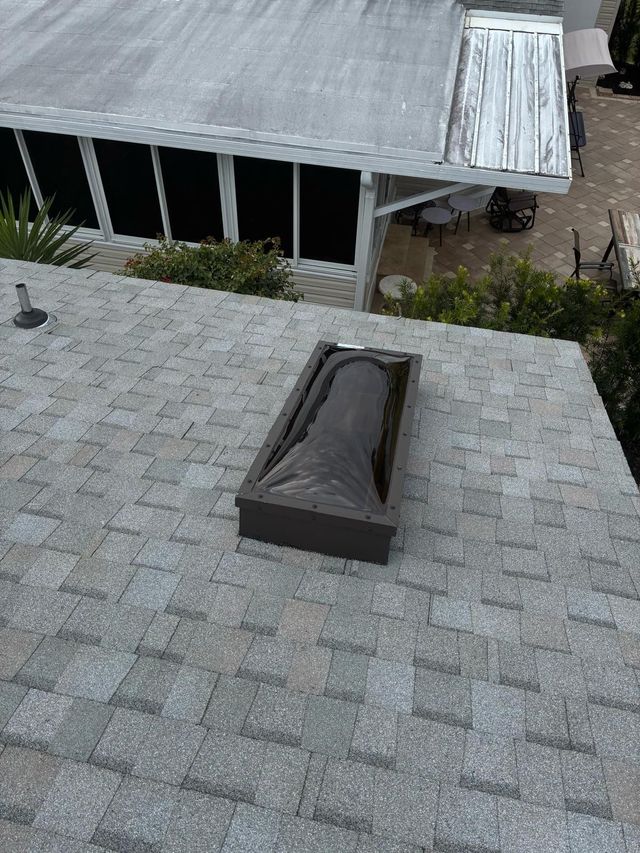 A rectangular skylight with a dark cover sits on a gray shingled roof, with a section of patio cover visible below.
