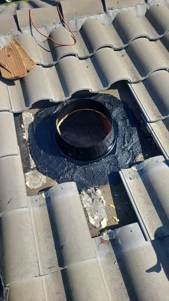 A circular roof vent surrounded by black flashing on a tile roof, with some surrounding roof tiles removed.