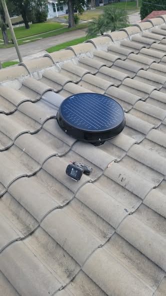 A solar-powered roof attic fan and a small electronic sensor are mounted on a concrete tile roof.
