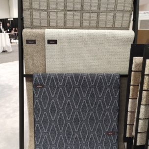 A display rack holding several carpet samples of varying textures and patterns in shades of gray, beige, and dark blue.