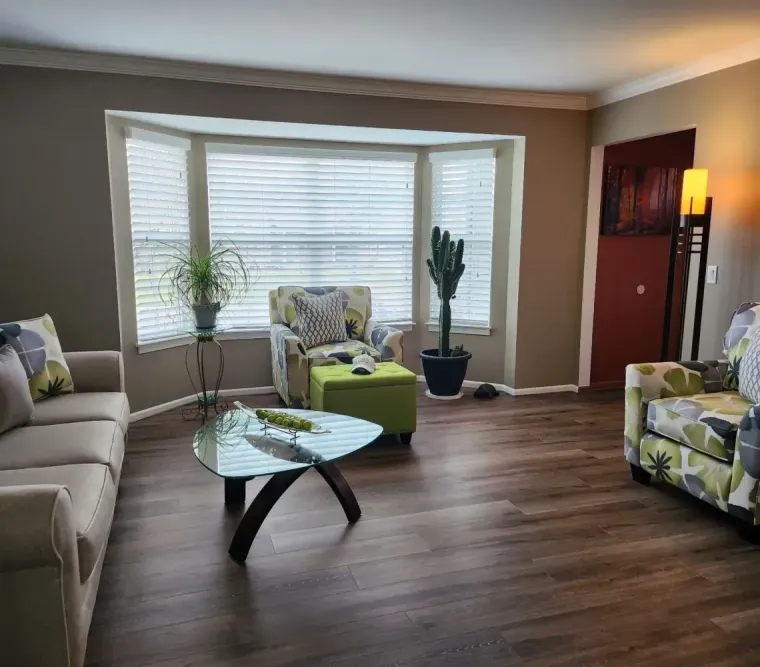 A living room with a beige sofa, a patterned armchair, a glass coffee table, a lime green ottoman, and a potted cactus.