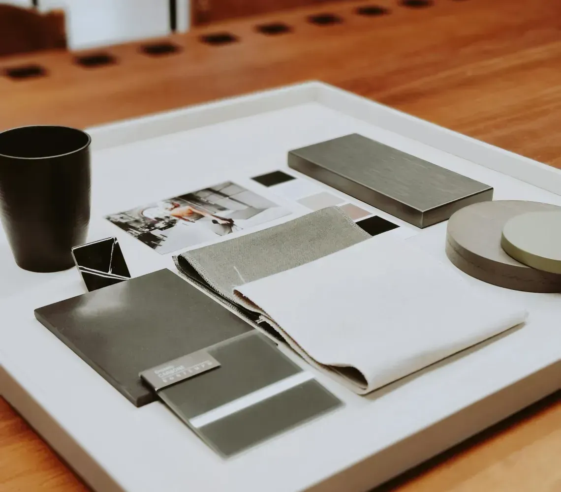 A white tray on a wooden table holds swatches of gray and white fabric, paint samples, and a black mug.