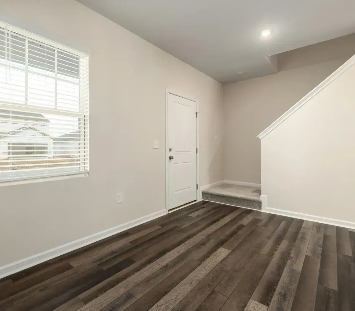 An interior view of a room with light beige walls, dark wood-look flooring, a window with blinds, and stairs.