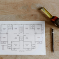 A top-down view of a blueprint on a wooden table, next to a coffee cup, a measuring tape, and a mechanical pencil.