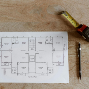 A top-down view of a blueprint on a wooden table, next to a coffee cup, a measuring tape, and a mechanical pencil.