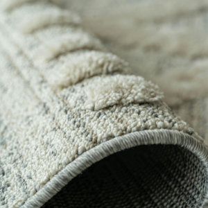 A close-up view of a rolled, textured beige and grey area rug showing the layered pile and woven fabric backing.