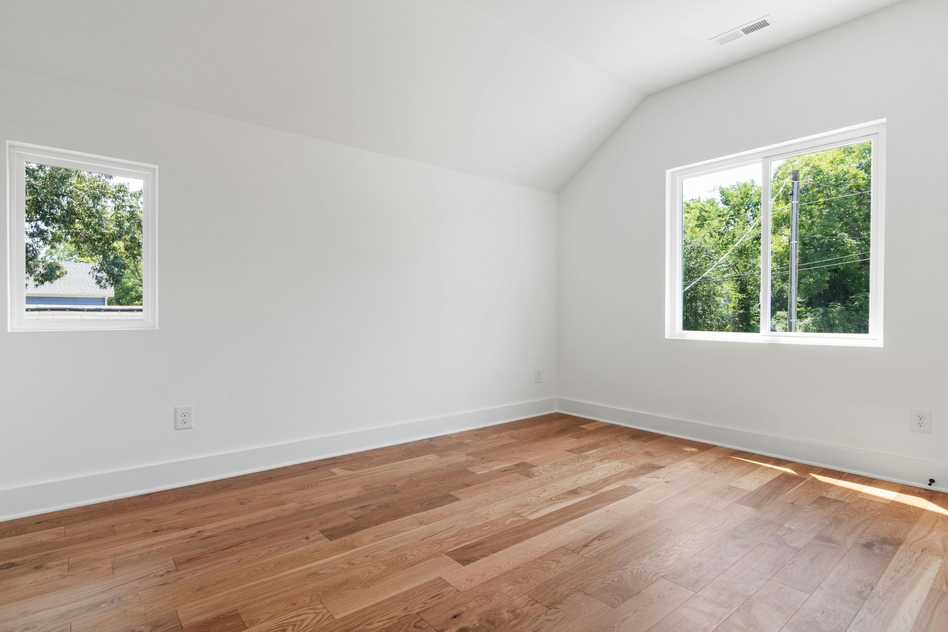 Empty room with white walls, slanted ceiling, hardwood floors, and two windows overlooking trees.