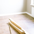Two sets of hands roll a textured, beige area rug on a wooden floor, securing the edge with a piece of blue painter's tape.