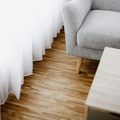 A grey armchair on light wood flooring sits next to white, floor-length curtains in a brightly lit room.