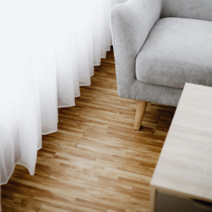 A grey armchair on light wood flooring sits next to white, floor-length curtains in a brightly lit room.