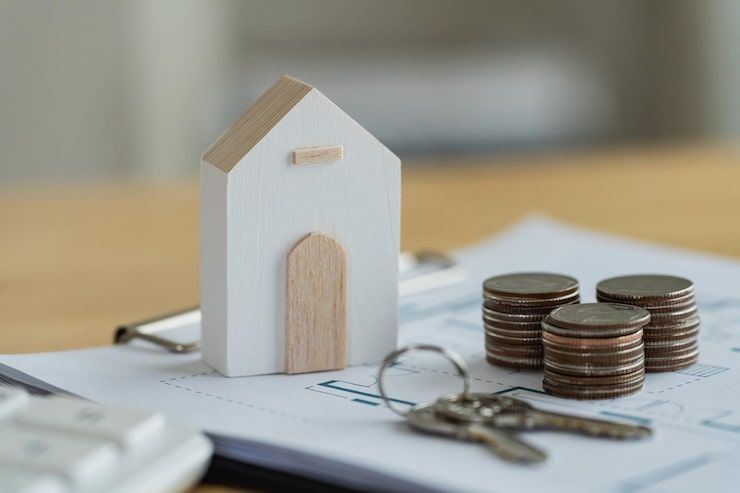 Mini house model beside stacked coins and keys on paperwork, suggesting home savings or mortgage planning