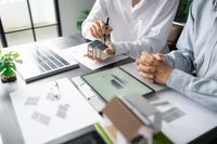 Two people review architectural model plans at a desk with a laptop and tablet