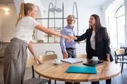 Three coworkers shake hands across a round table in a bright office with laptops and papers.