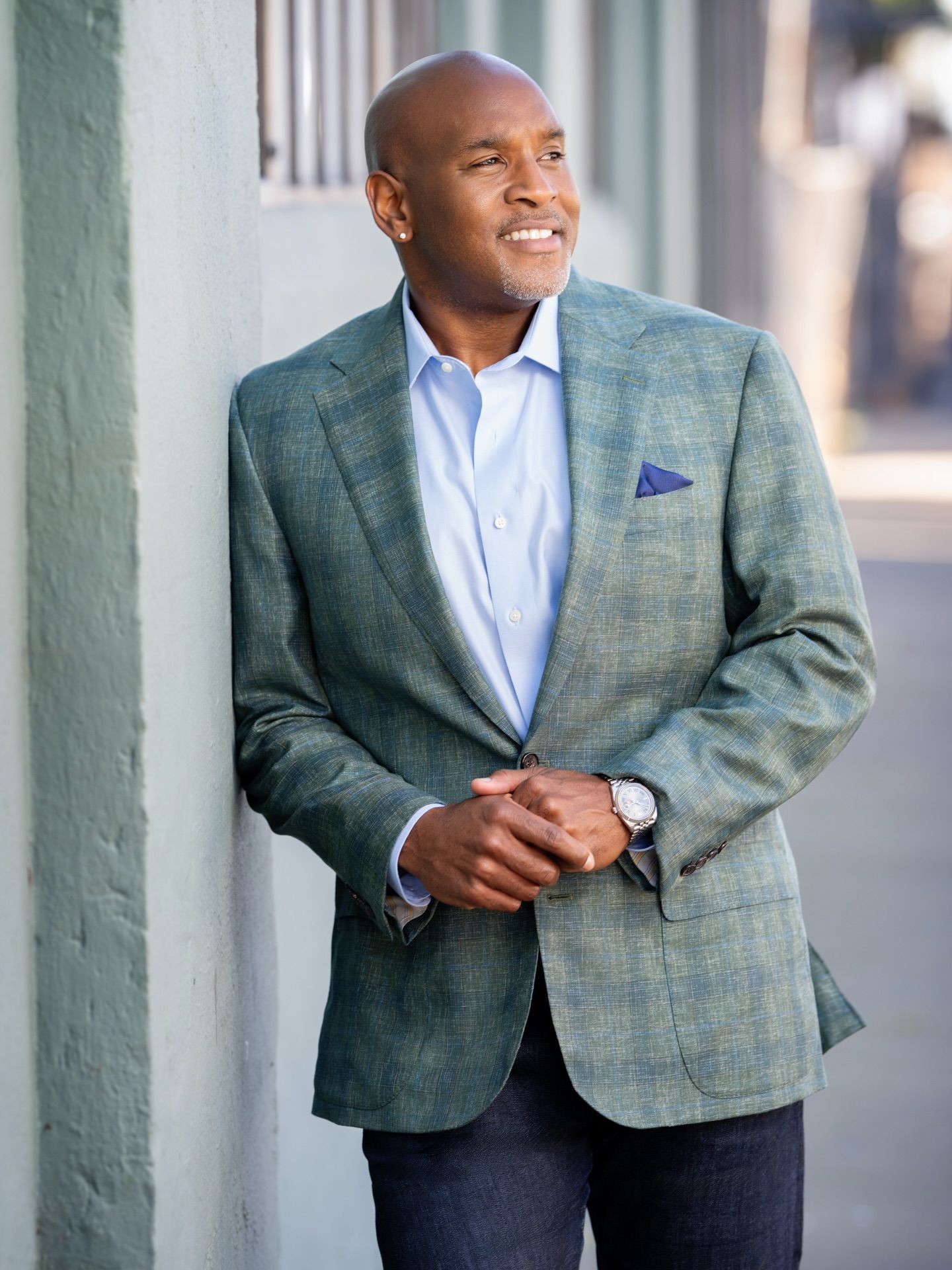 Man in a green blazer leaning against a wall, looking off to the side with a slight smile.