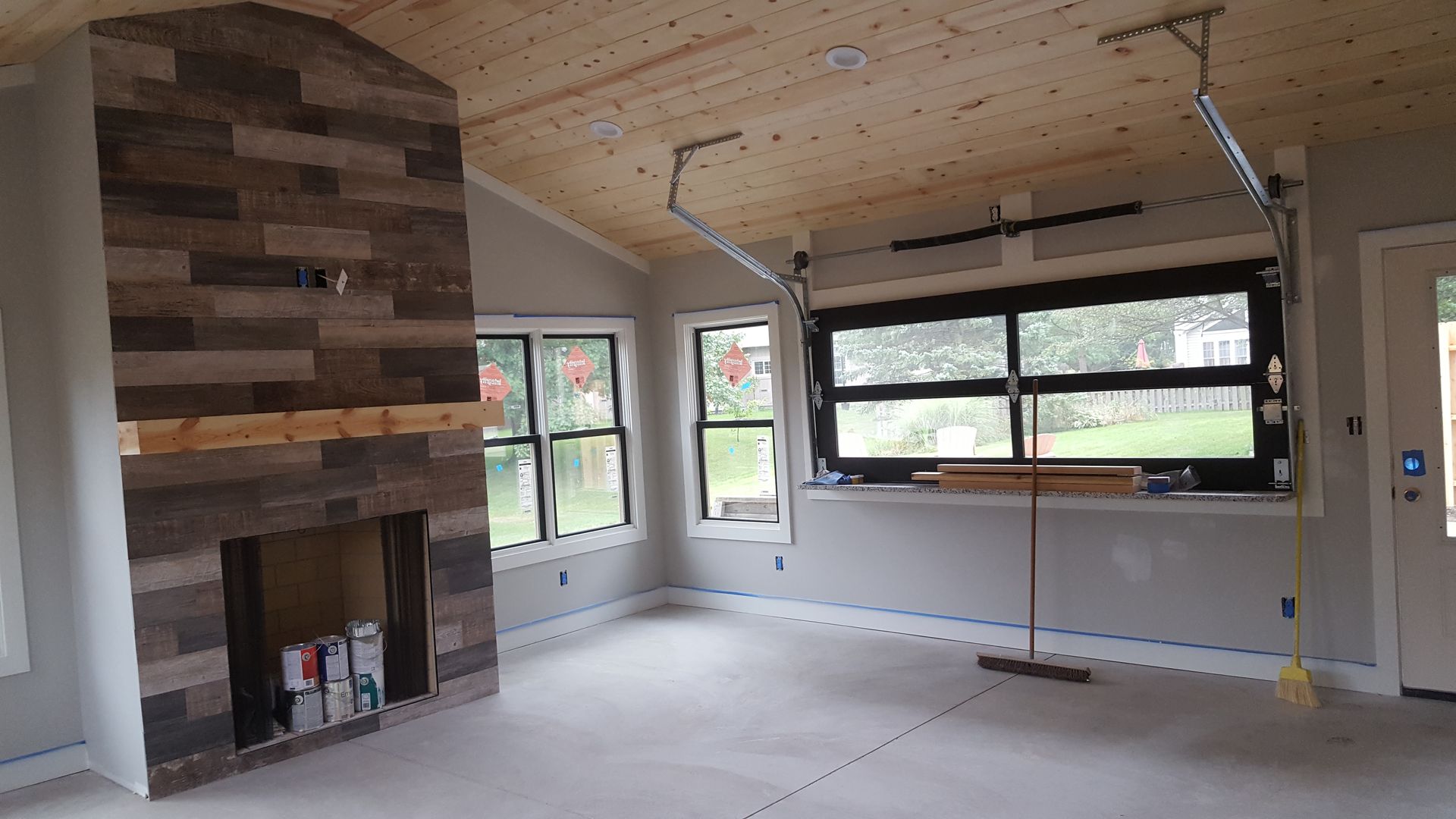 Empty living room with unfinished fireplace, large windows, and bare floors under renovation