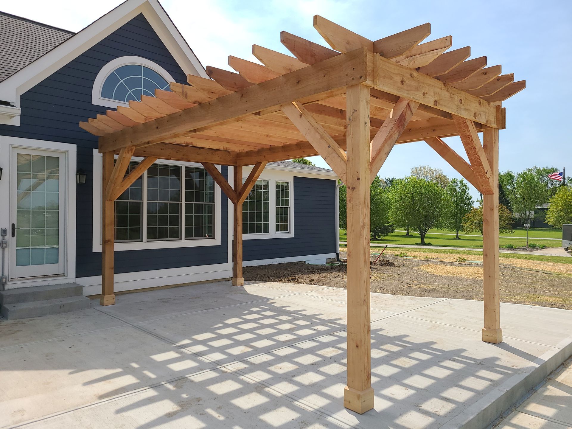 Wooden pergola casting striped shadows on a concrete patio beside a dark blue house