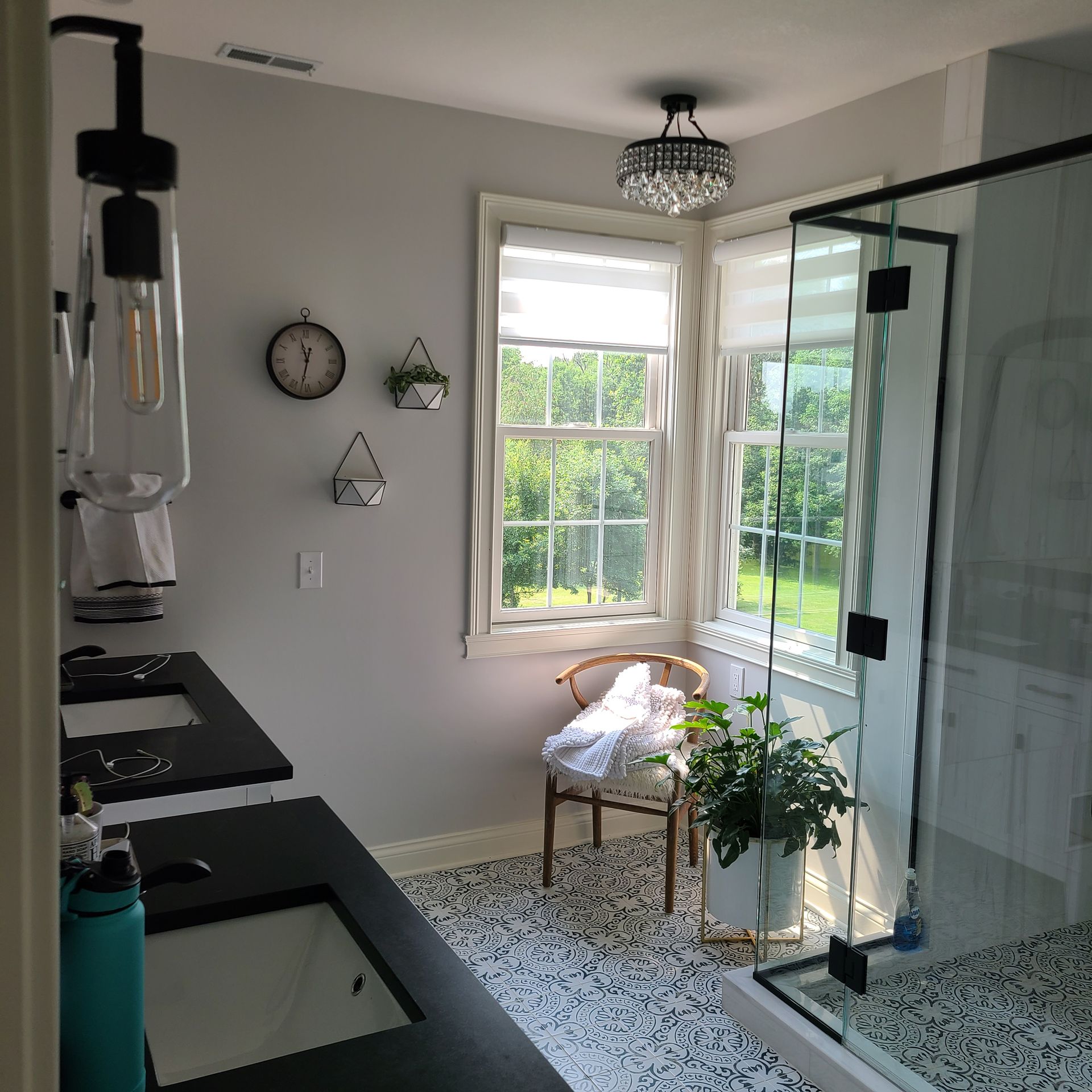 Bright bathroom with black fixtures, glass shower, patterned tile, and a chair by the window.