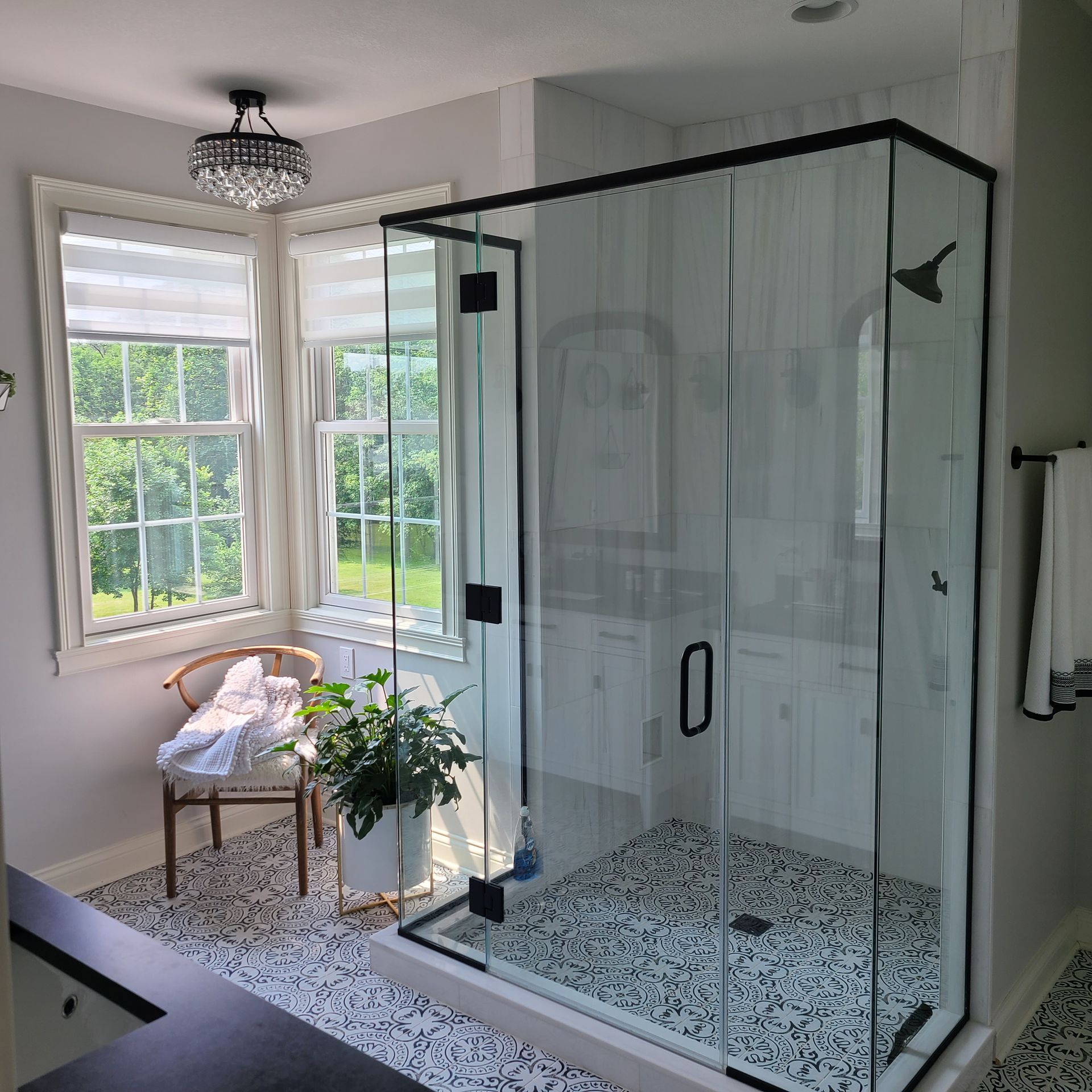 Bright bathroom with a glass shower, patterned tile floor, and a chair by large windows.