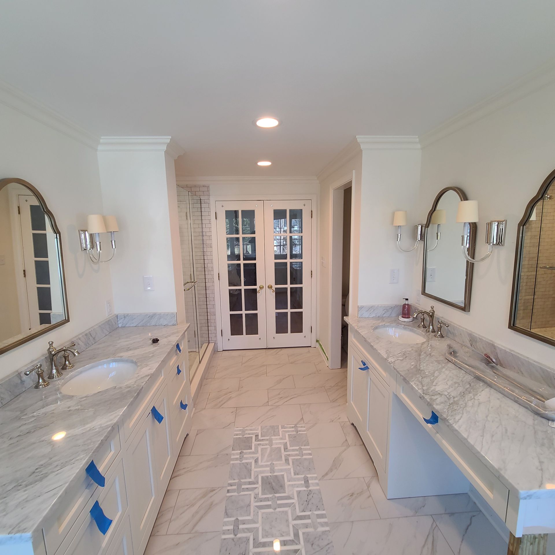 Bright white double vanity bathroom with marble counters, mirrors, sconces, and French doors at the end
