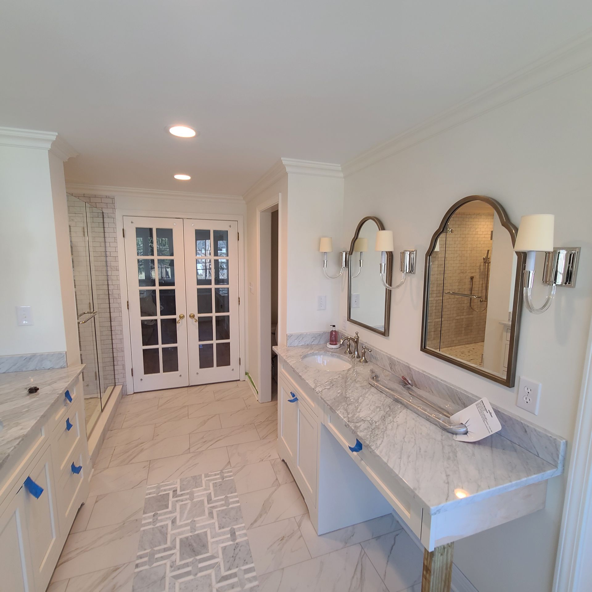 Bright bathroom with double vanity, marble countertops, mirrors, and French doors at the end