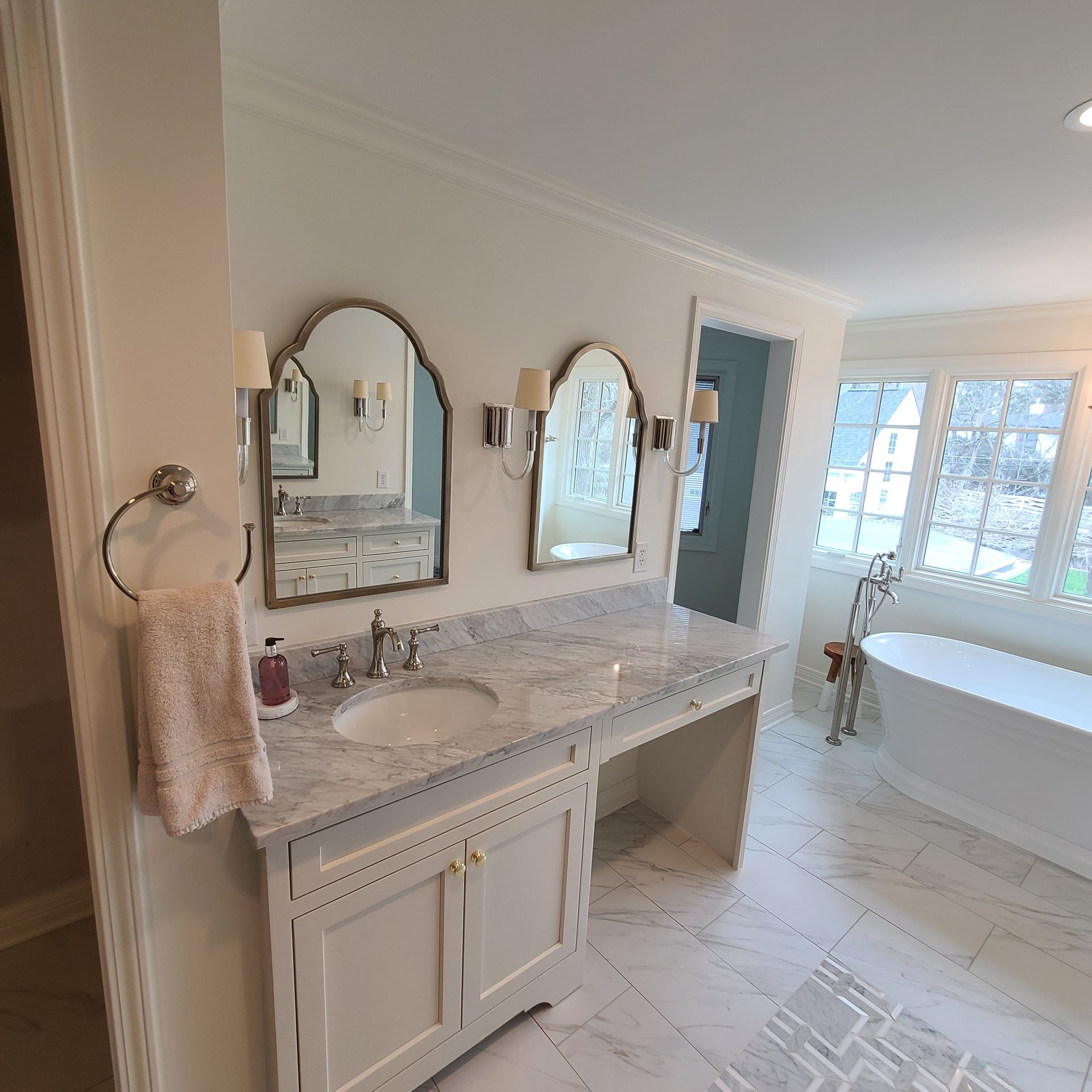 Bright white bathroom with double vanity, marble countertop, mirrors, and a freestanding tub by windows.