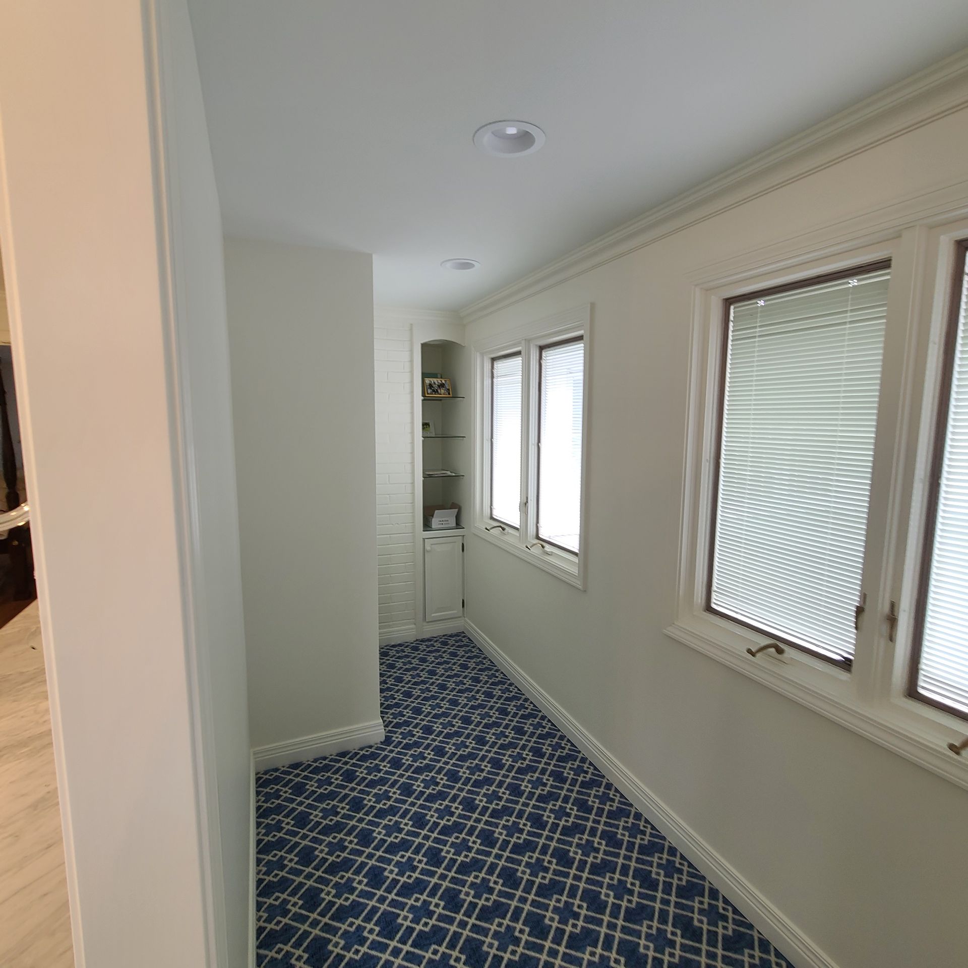Narrow hallway with white walls, patterned blue carpet, and windows along the right side.