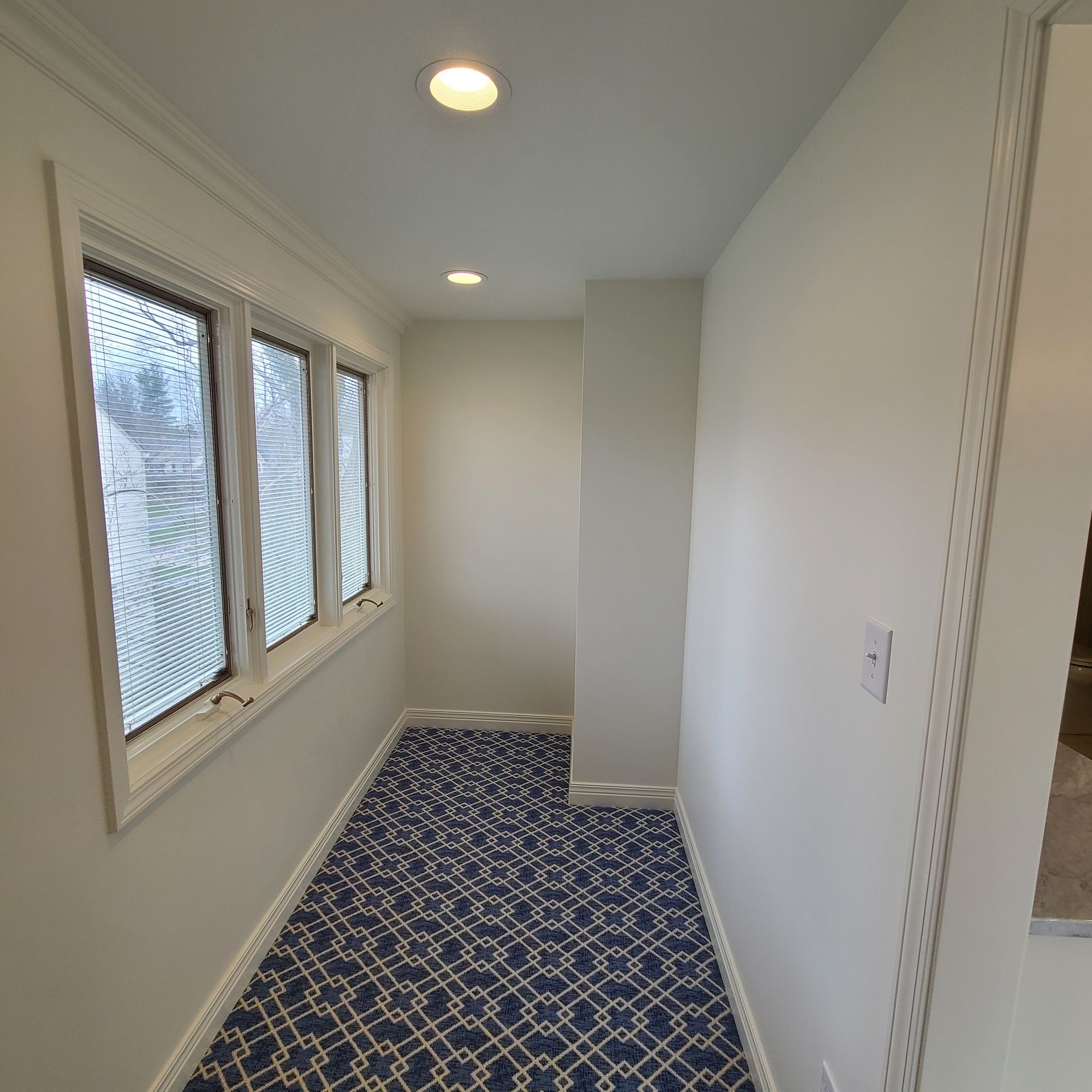 Narrow carpeted hallway with white walls, recessed lights, and windows along the left side.