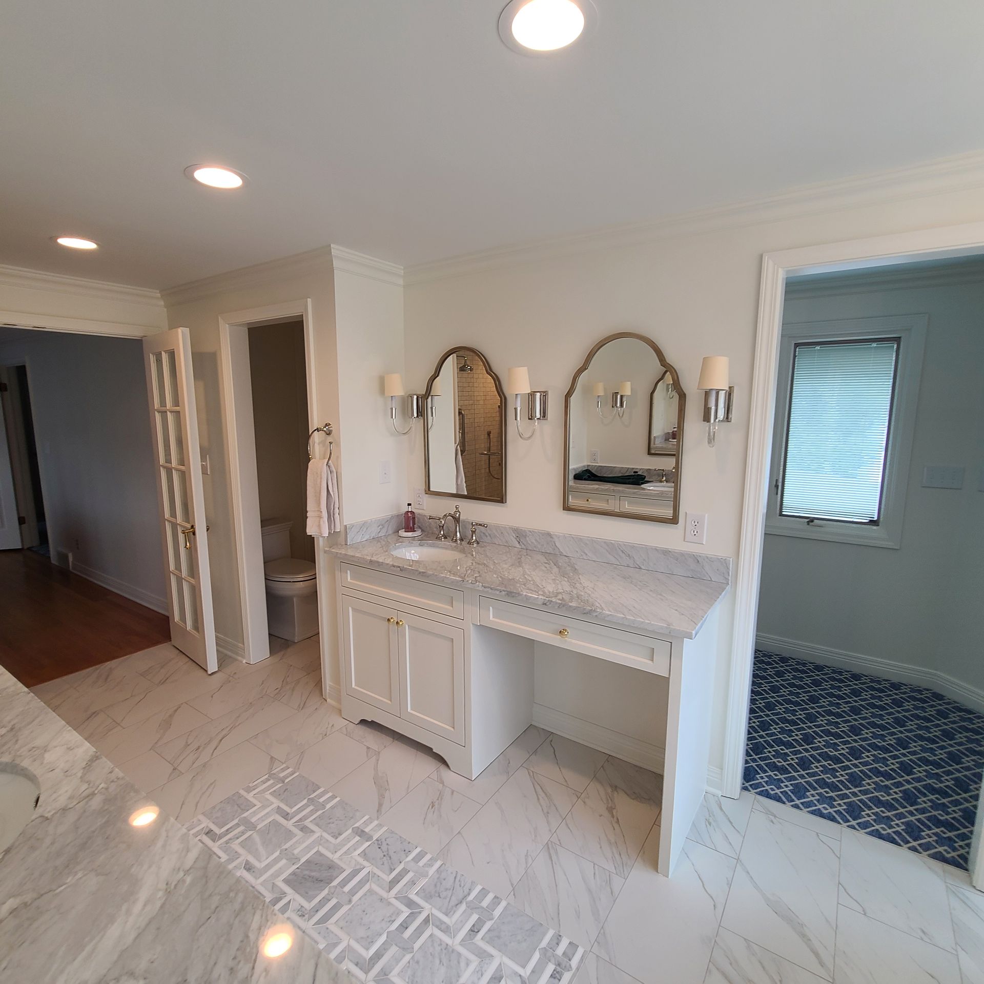 Bright bathroom with marble vanity, twin mirrors, white cabinets, and blue-tiled adjacent room.