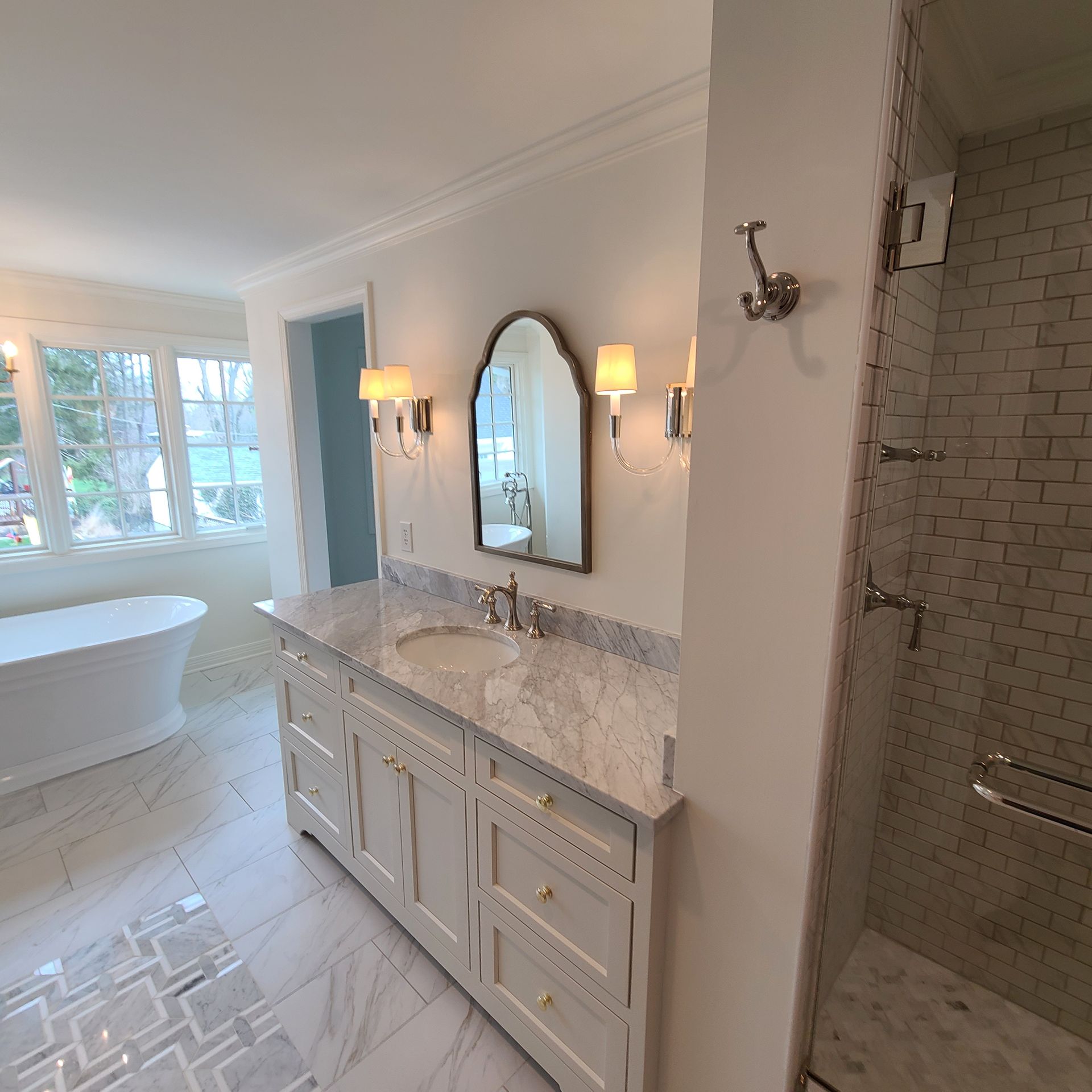 Bright bathroom with white tub, marble vanity, arched mirror, and glass shower.