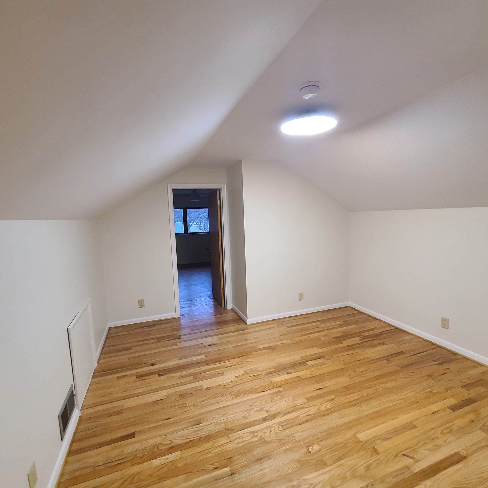 Empty attic room with sloped white ceiling, hardwood floor, and a ceiling light.