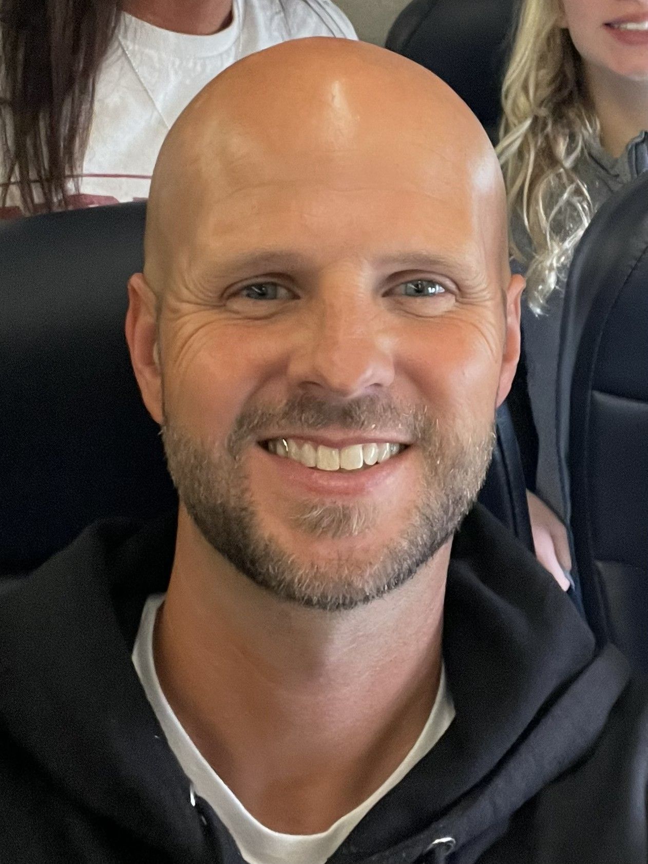 Smiling bald man in a black hoodie seated in a car, with two people blurred behind him.