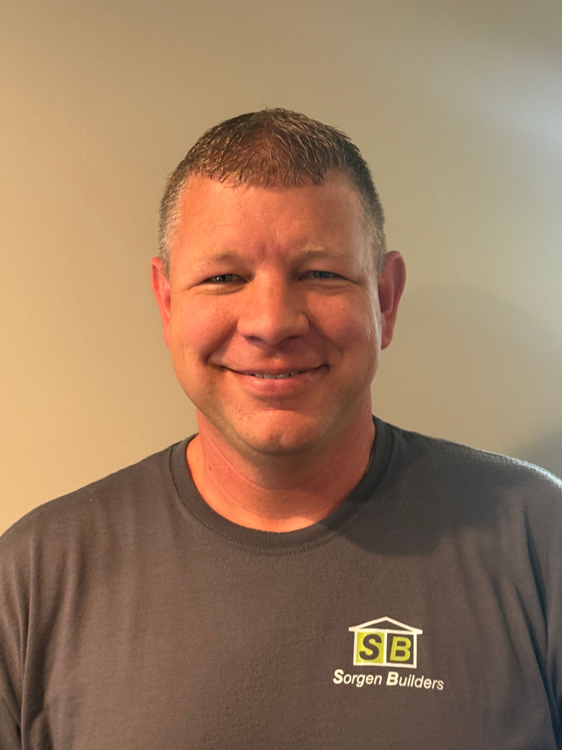 Smiling man in a gray T-shirt with a small logo, standing against a plain beige wall.