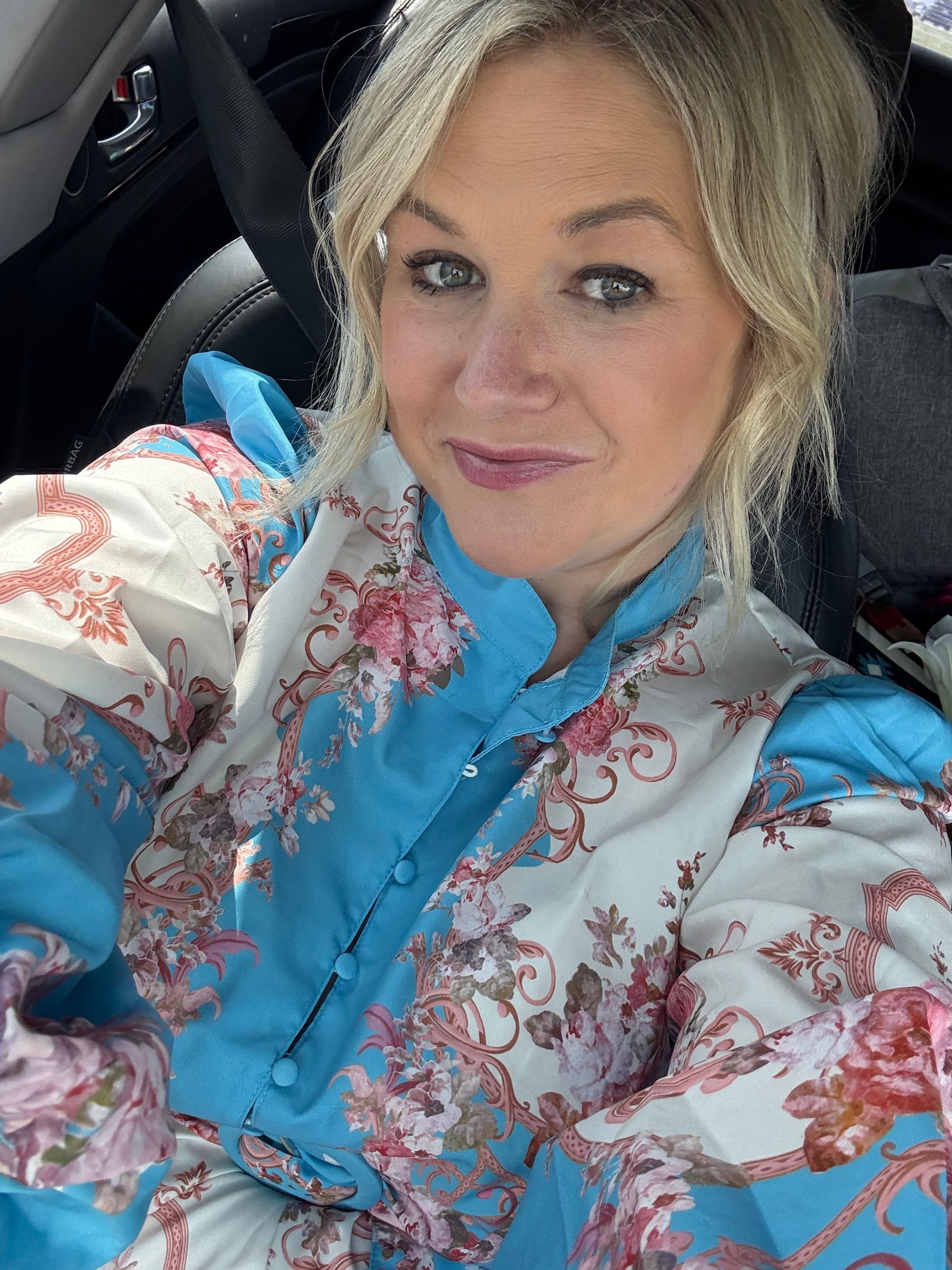 Woman in a car, wearing a blue blouse and floral jacket, looking at the camera and smiling.