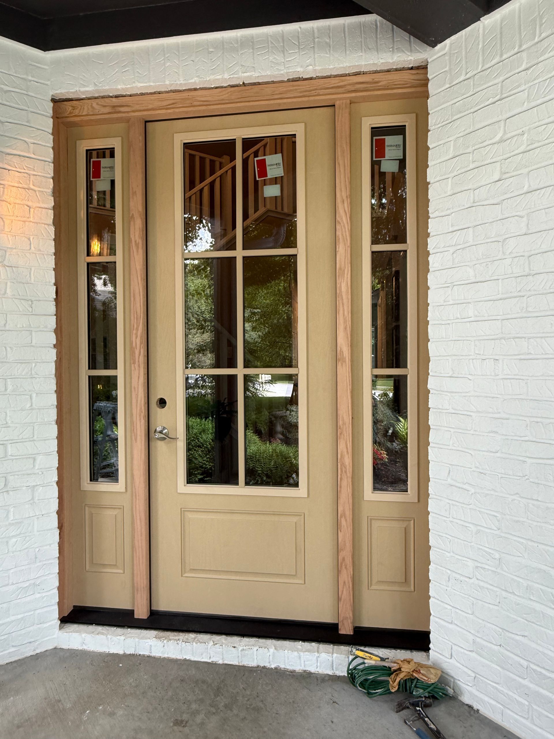 Beige front door with glass panels and sidelights under a covered porch against a white brick wall