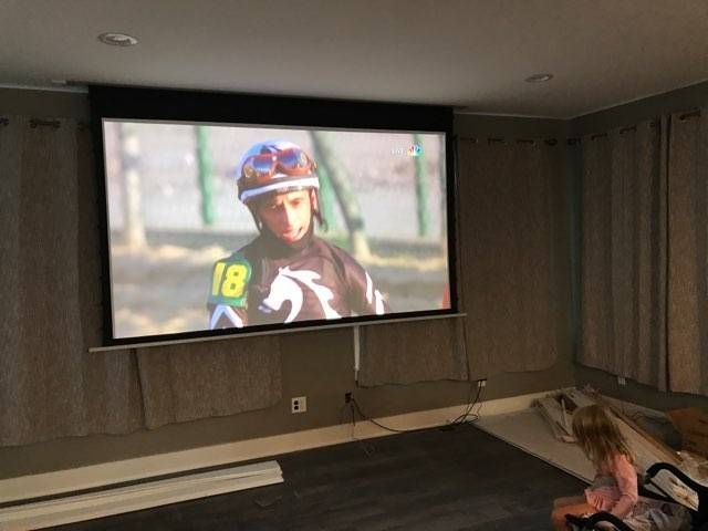 Home theater with a projected horse racing scene on a large screen and a person sitting on the floor at right.