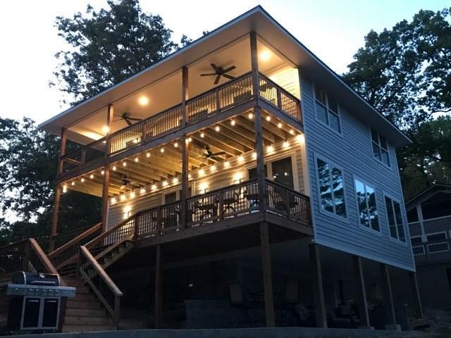 Two-story wooden house with lit wraparound porch at dusk, on stilts among trees