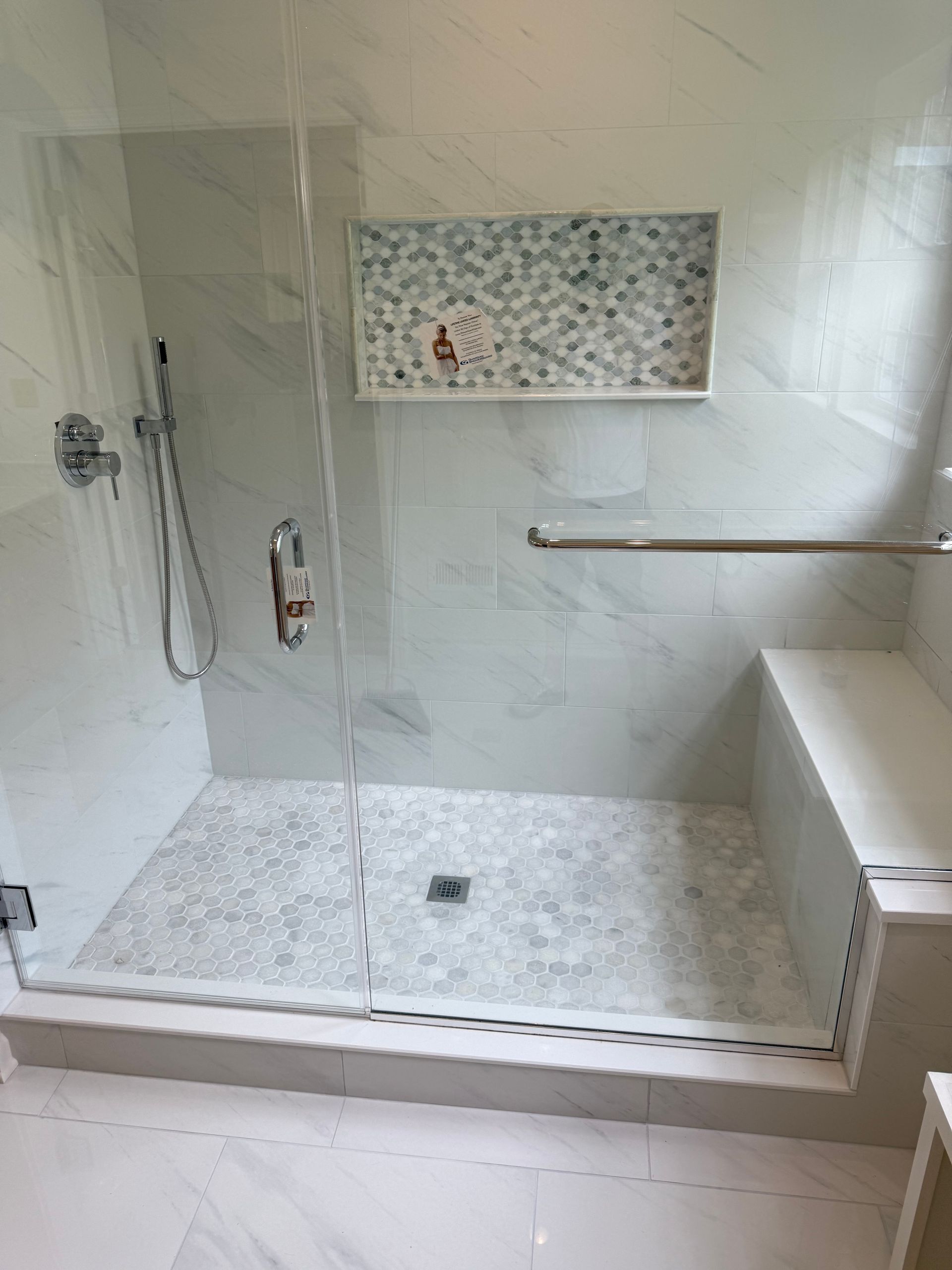 Modern white tiled walk-in shower with glass door, handheld showerhead, grab bar, and bench seat