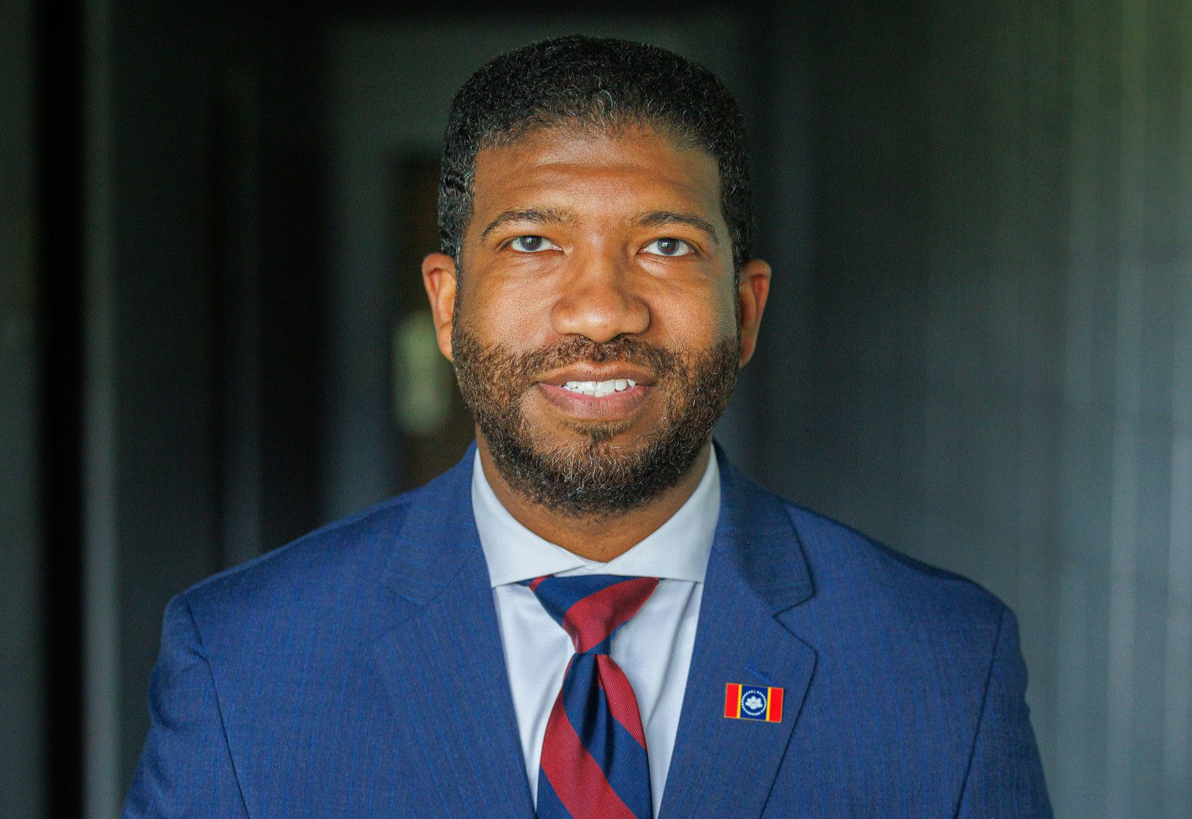 A man in a blue suit and red tie is sitting at a table with his hands folded.