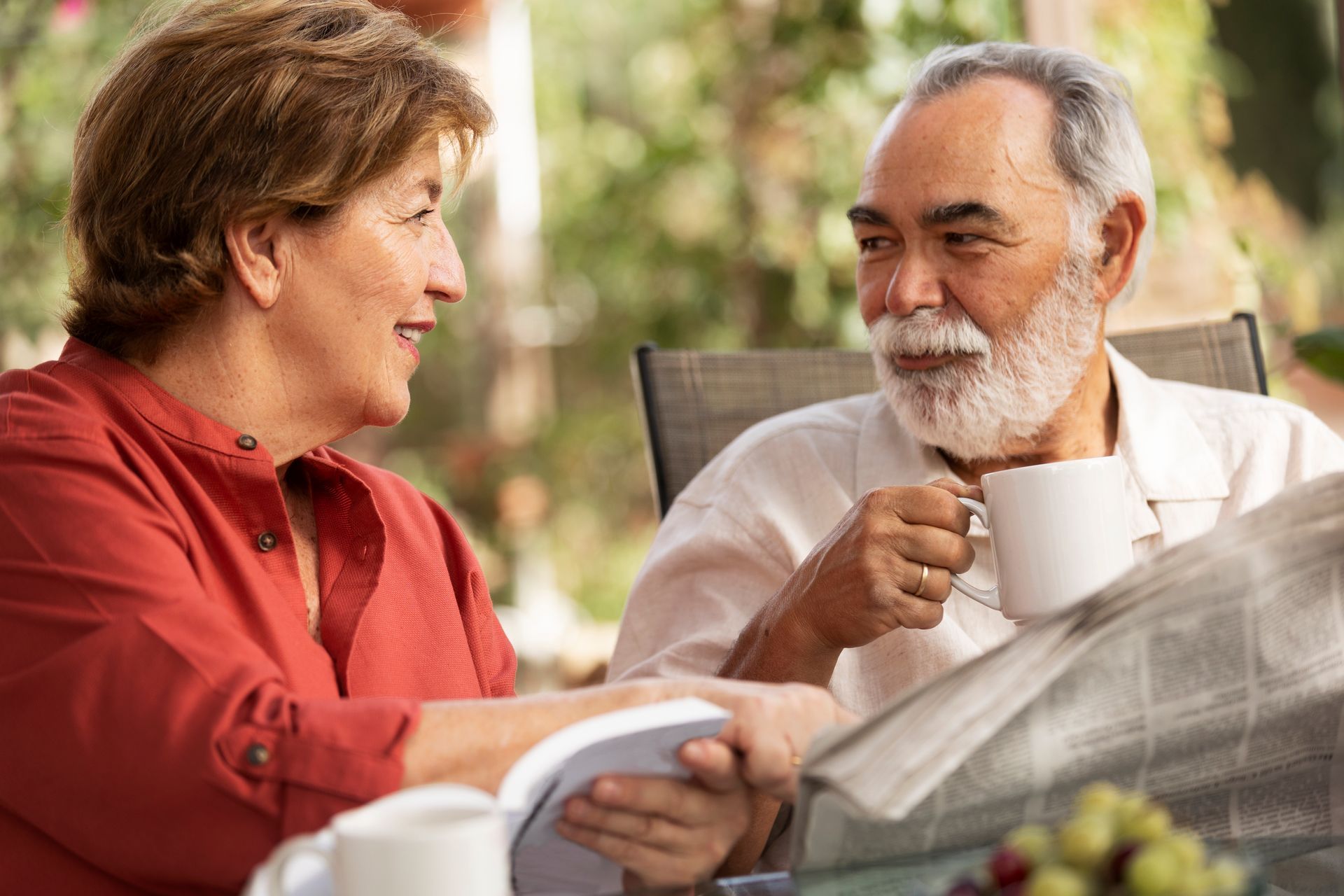 An elderly couple is sitting at a table drinking coffee and reading a newspaper.