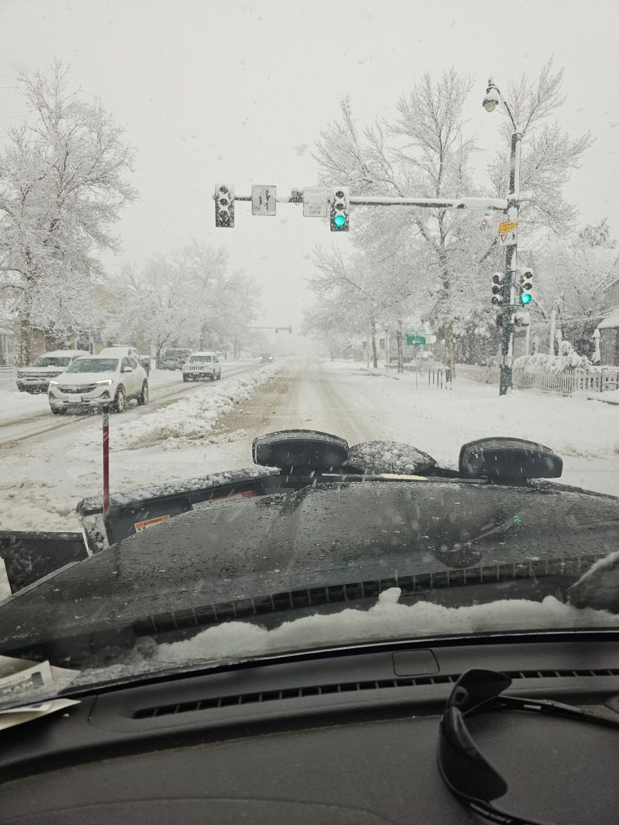 Snowy street scene, viewed from inside a vehicle. Traffic signals lit, vehicles driving on snow-covered road.