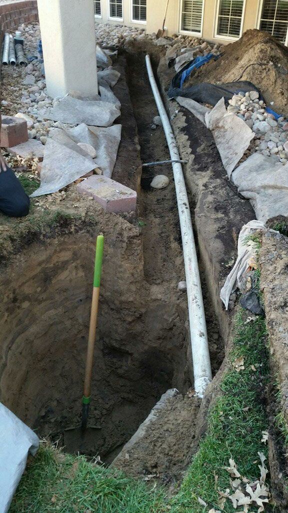 A trench dug near a building with a white pipe running through it. A shovel leans against the side.