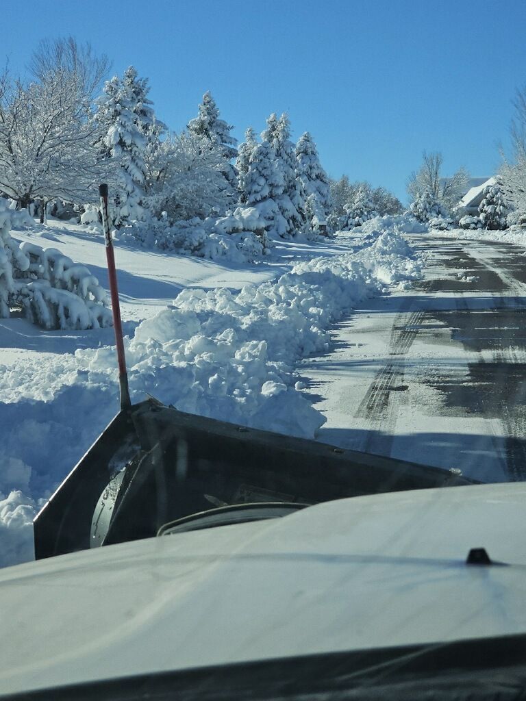 Snowplow clearing a snow-covered road on a sunny day. Trees and snowdrifts line the road.
