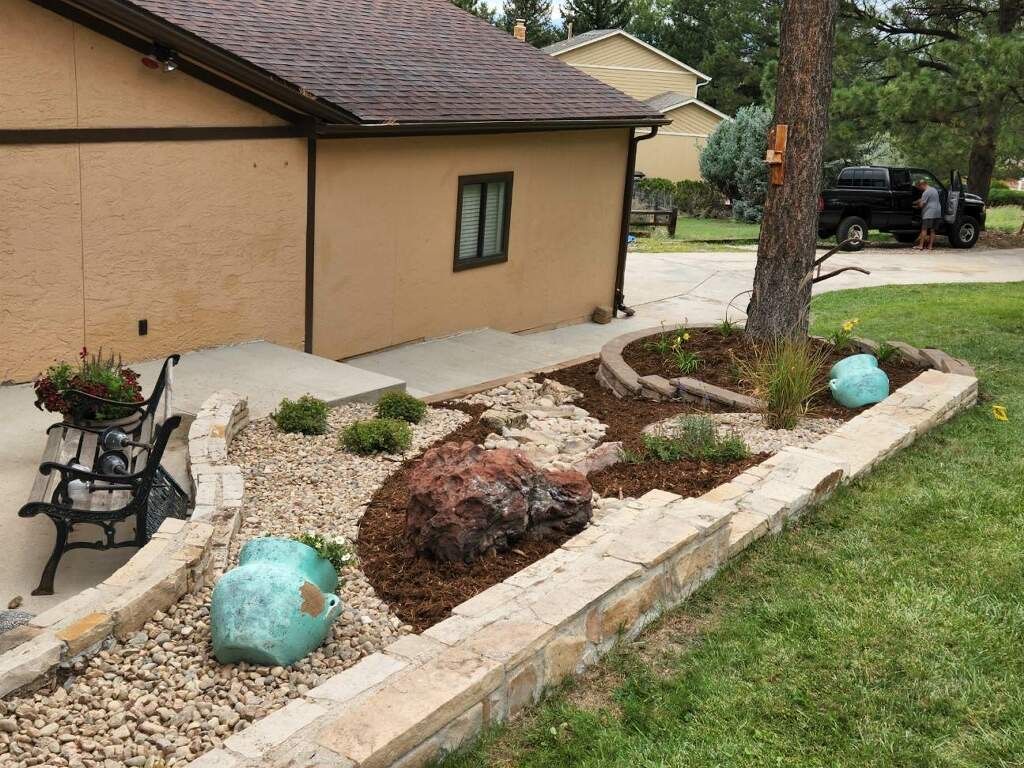 Landscaped yard with stone retaining wall, mulch, rocks, and a bench in front of a tan house.