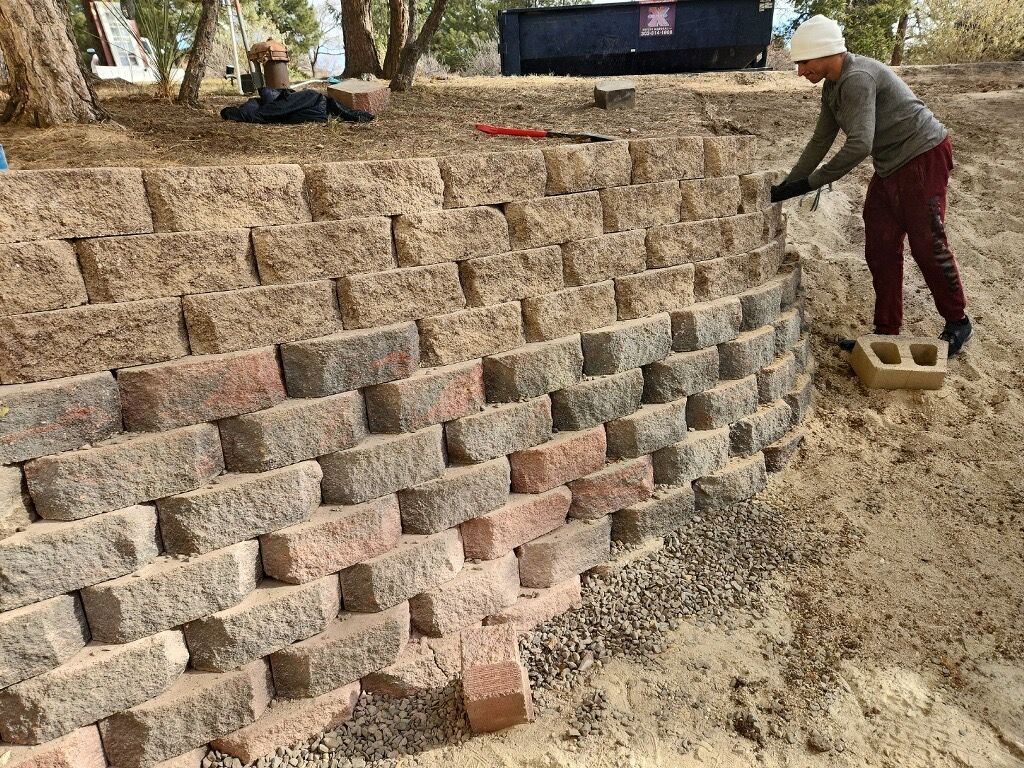 Person building a stone retaining wall, using blocks of varying colors, outdoors.
